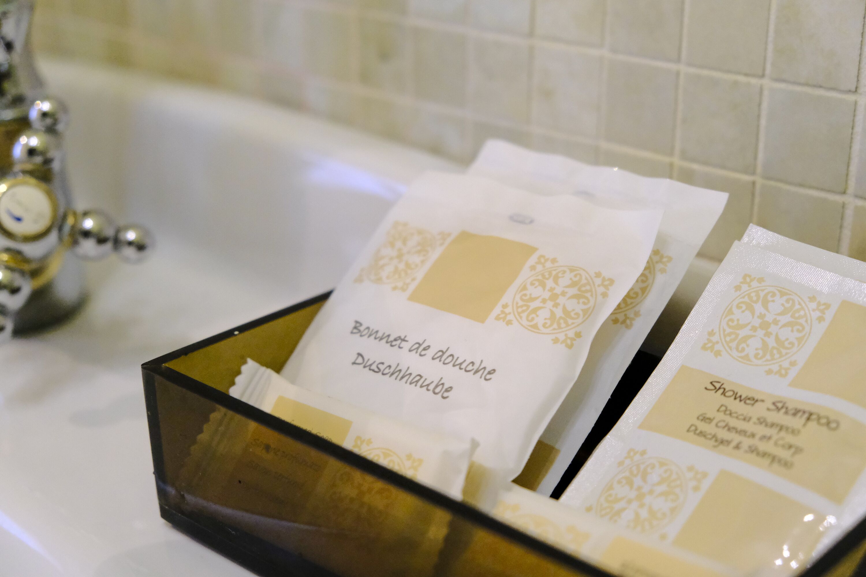 A bathroom with a glass container holding two toiletry bags labeled "Bonnet de douche" and "Shower Shampoo".