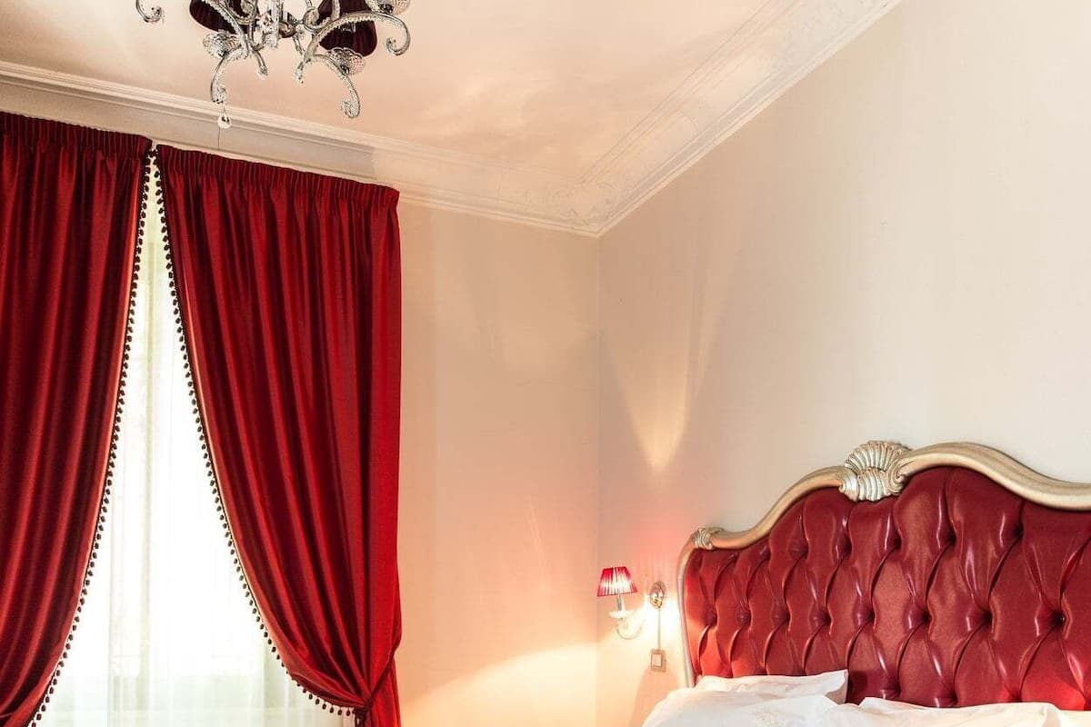 Ein Hotelzimmer mit einem großen Bett, roten Vorhängen, einer Kristallleuchtre und einem roten Kopfteil.