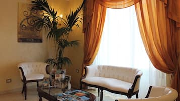 A living room with two white upholstered sofas, a coffee table with magazines, a potted plant, and a large window with curtains.