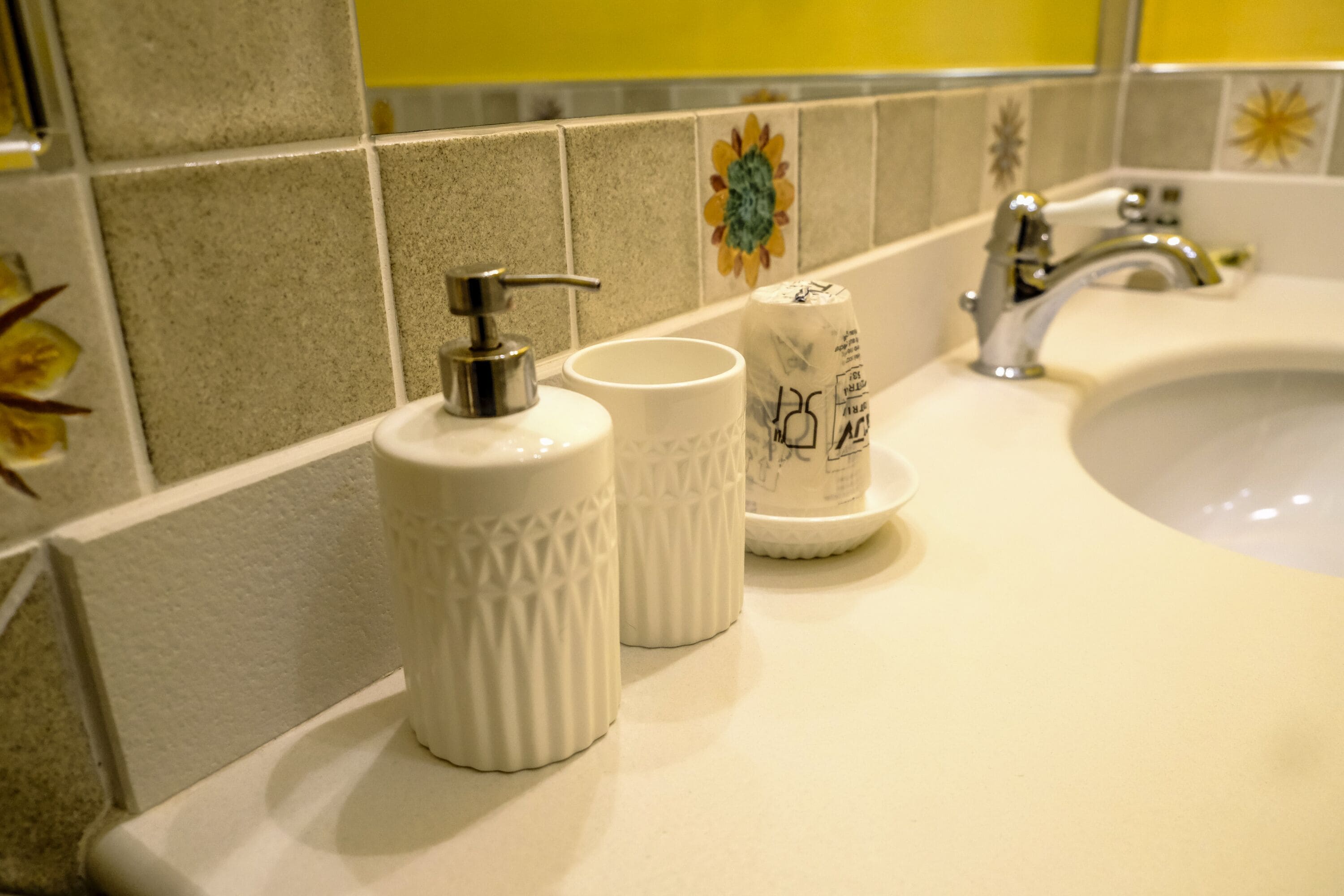 A bathroom sink with a white countertop, a silver faucet, and two white ceramic cups with a pump dispenser.