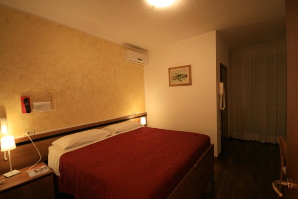 Una habitación de hotel con una cama individual, una mesita de noche con una lámpara, un equipo de aire acondicionado, un cuadro enmarcado en la pared y una puerta que da a otra habitación.