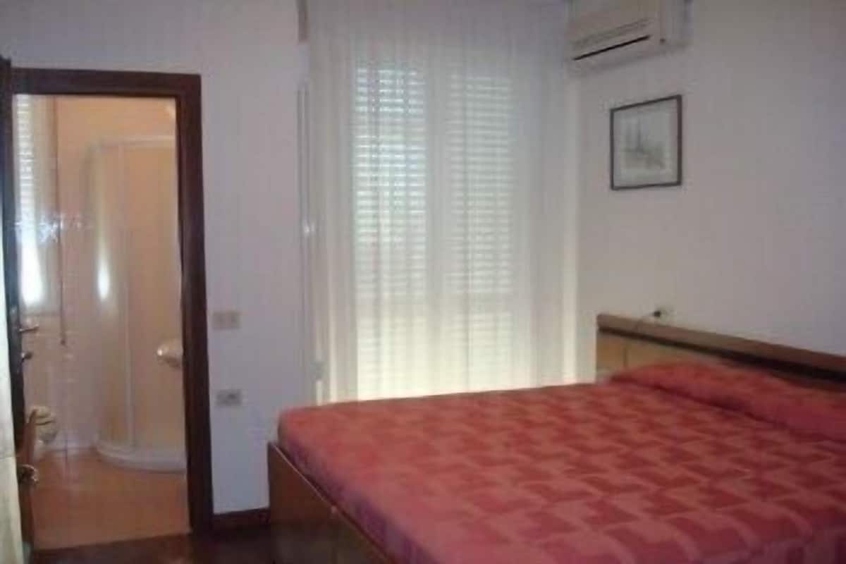 A bedroom with a bed, a door leading to another room, and a window with blinds.