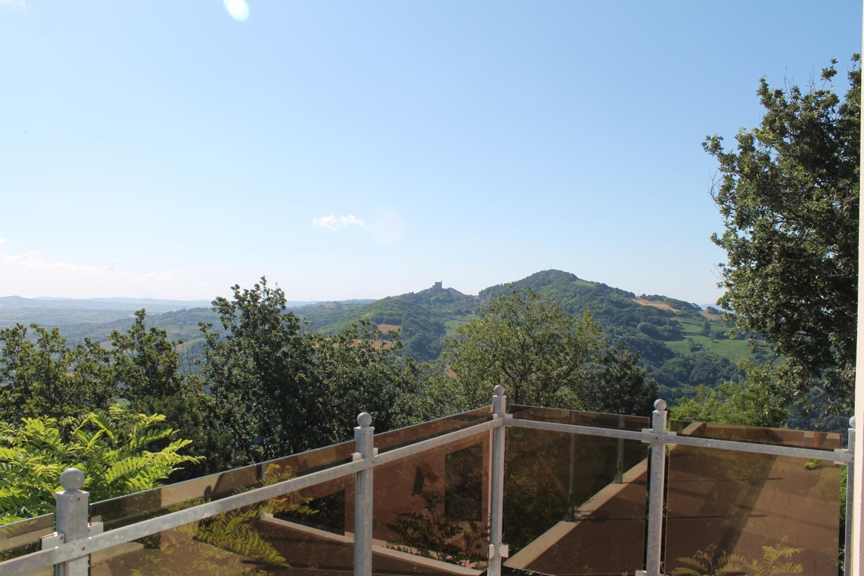A panoramic view from a balcony with a metal railing, overlooking a hilly landscape with dense greenery and distant hills.