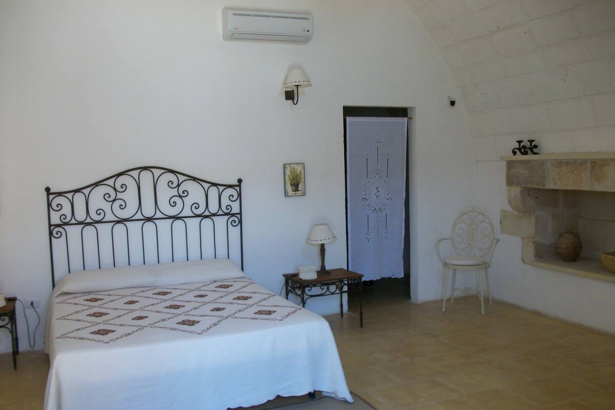 A bedroom with a bed, a nightstand, a chair, a wall-mounted lamp, a door, and a shelf with a vase.