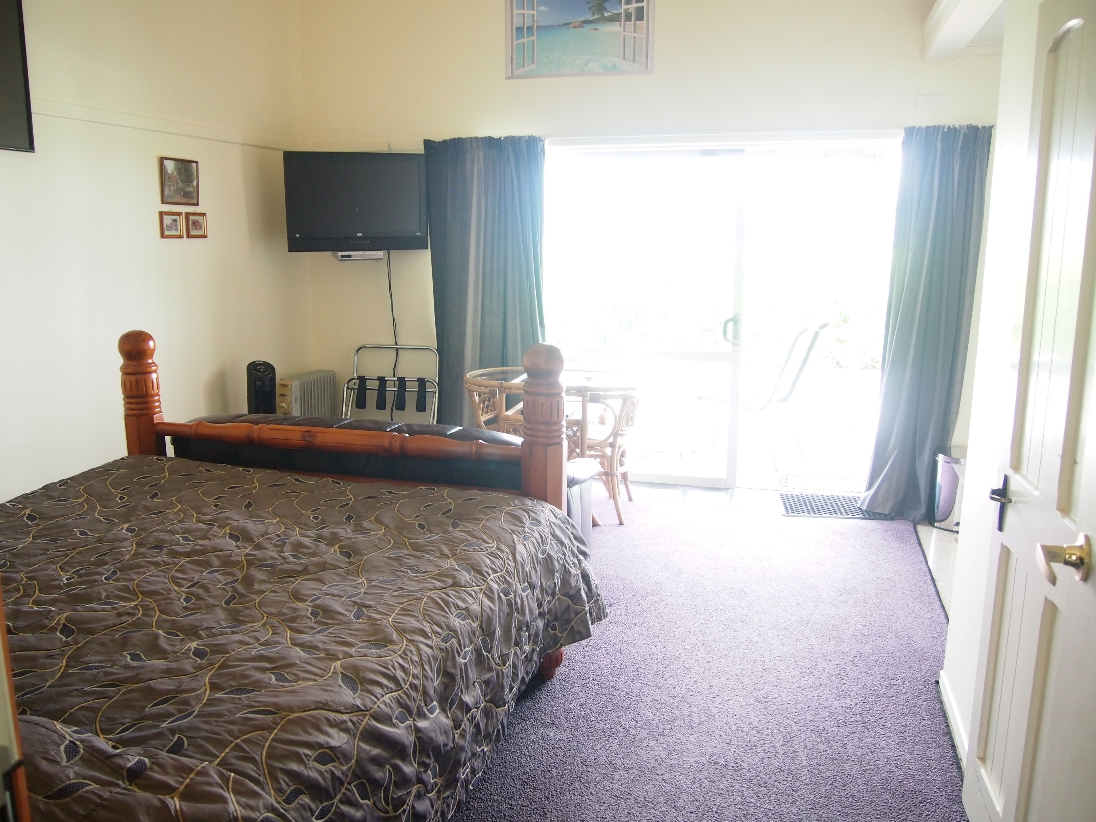 A hotel room with a bed, a television mounted on the wall, a small table with chairs, and a view of the outdoors through a window.