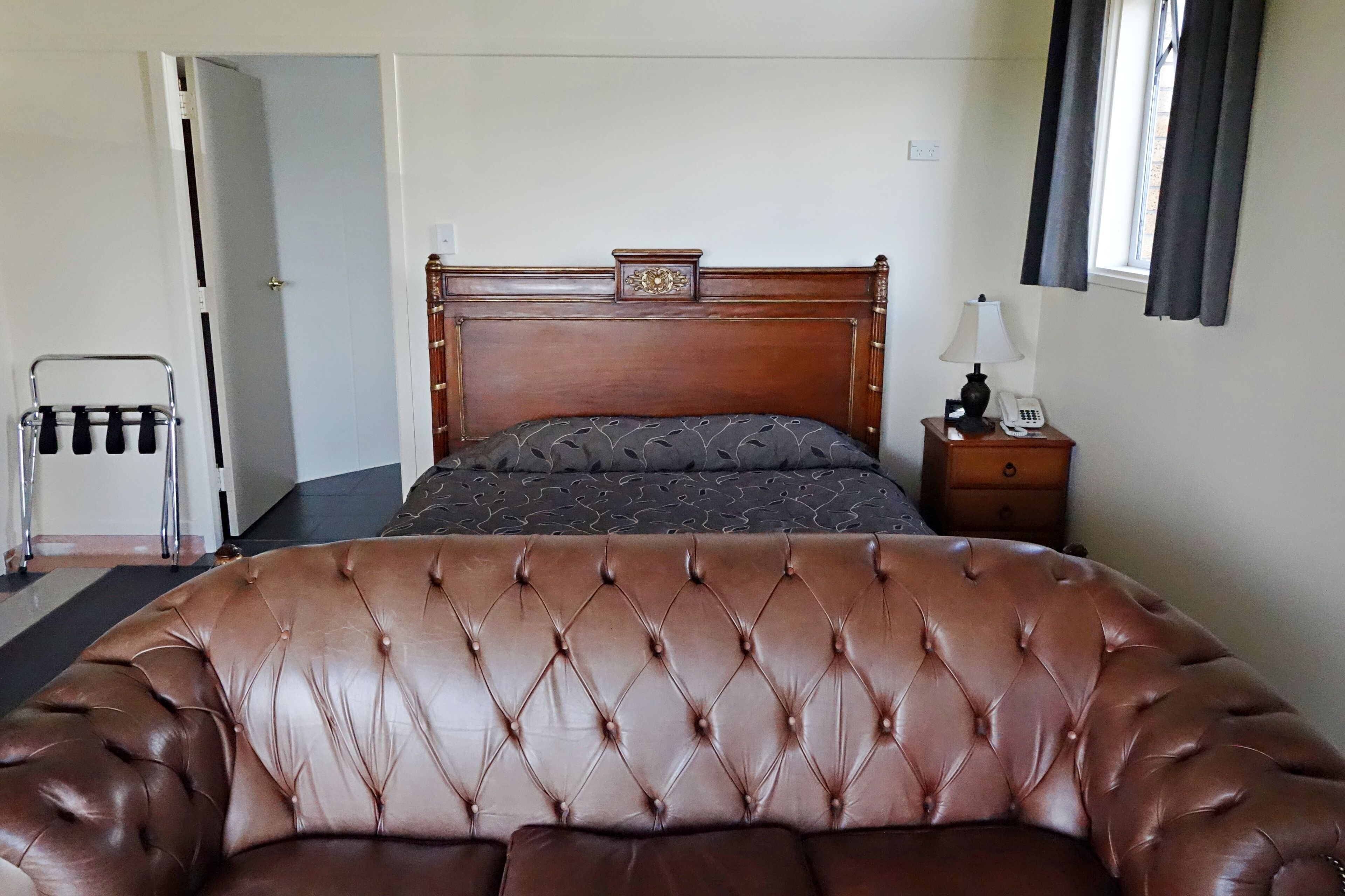 Un dormitorio con un amplio sofá de cuero, una cama de madera con cabecera, una mesita de noche con lámpara y una ventana con cortinas.