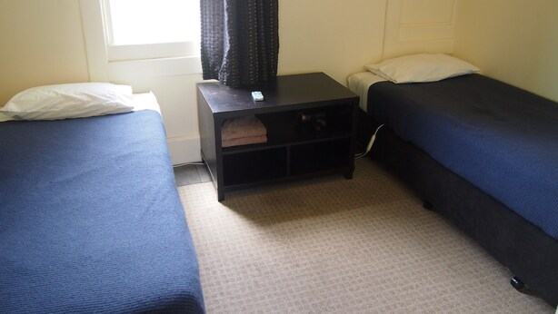 A room with two single beds, a small bedside table with a remote control, a power outlet on the wall, and a window with curtains. - Manor Views (Huntly)