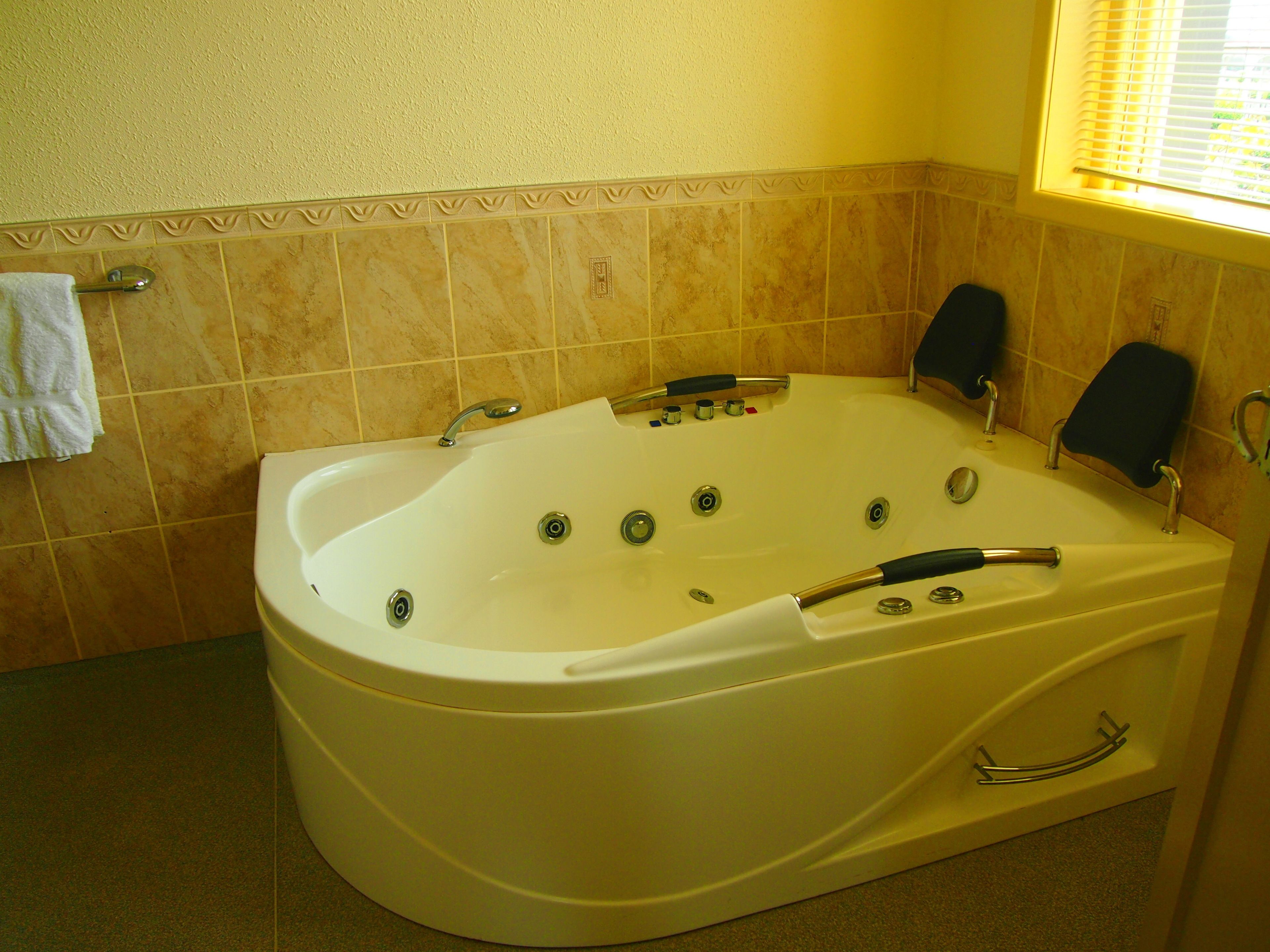 A large, freestanding bathtub in a bathroom with beige tiles, a towel rack, and a window with blinds.