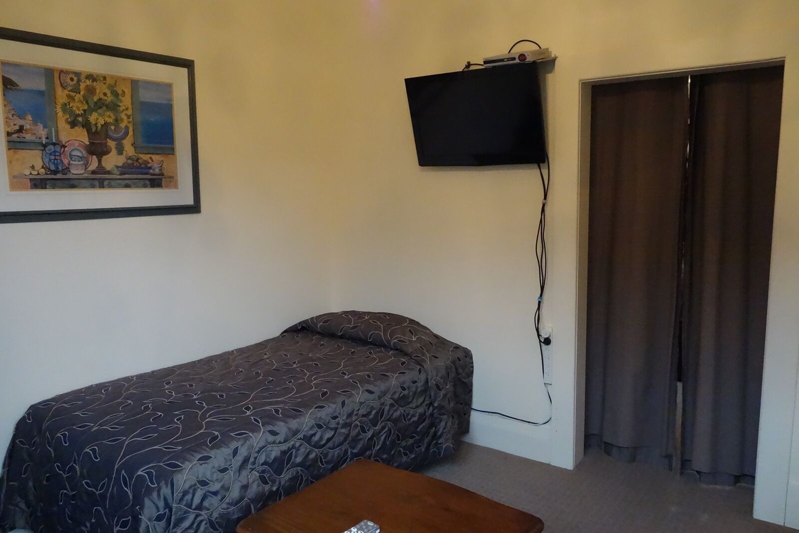 Una habitación pequeña con una cama, una mesa, un televisor montado en la pared y una puerta con cortinas.