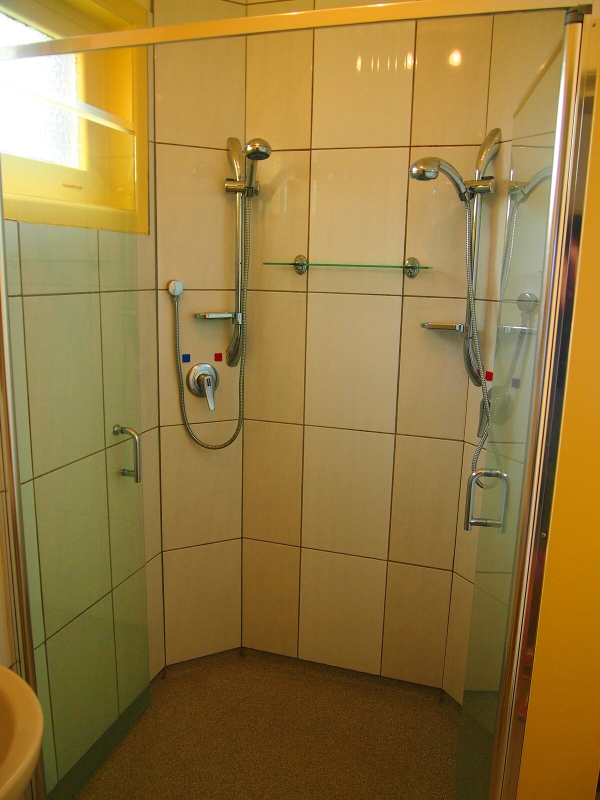 Chuveiro de canto com porta de vidro, chuveiro manual e torneira fixada na parede.