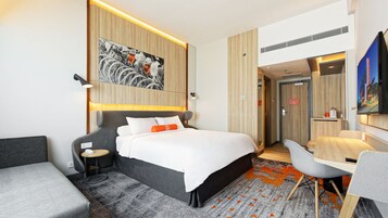 Habitación de hotel moderna con cama, escritorio y silla. Hay un cuadro en la pared y un televisor montado allí.