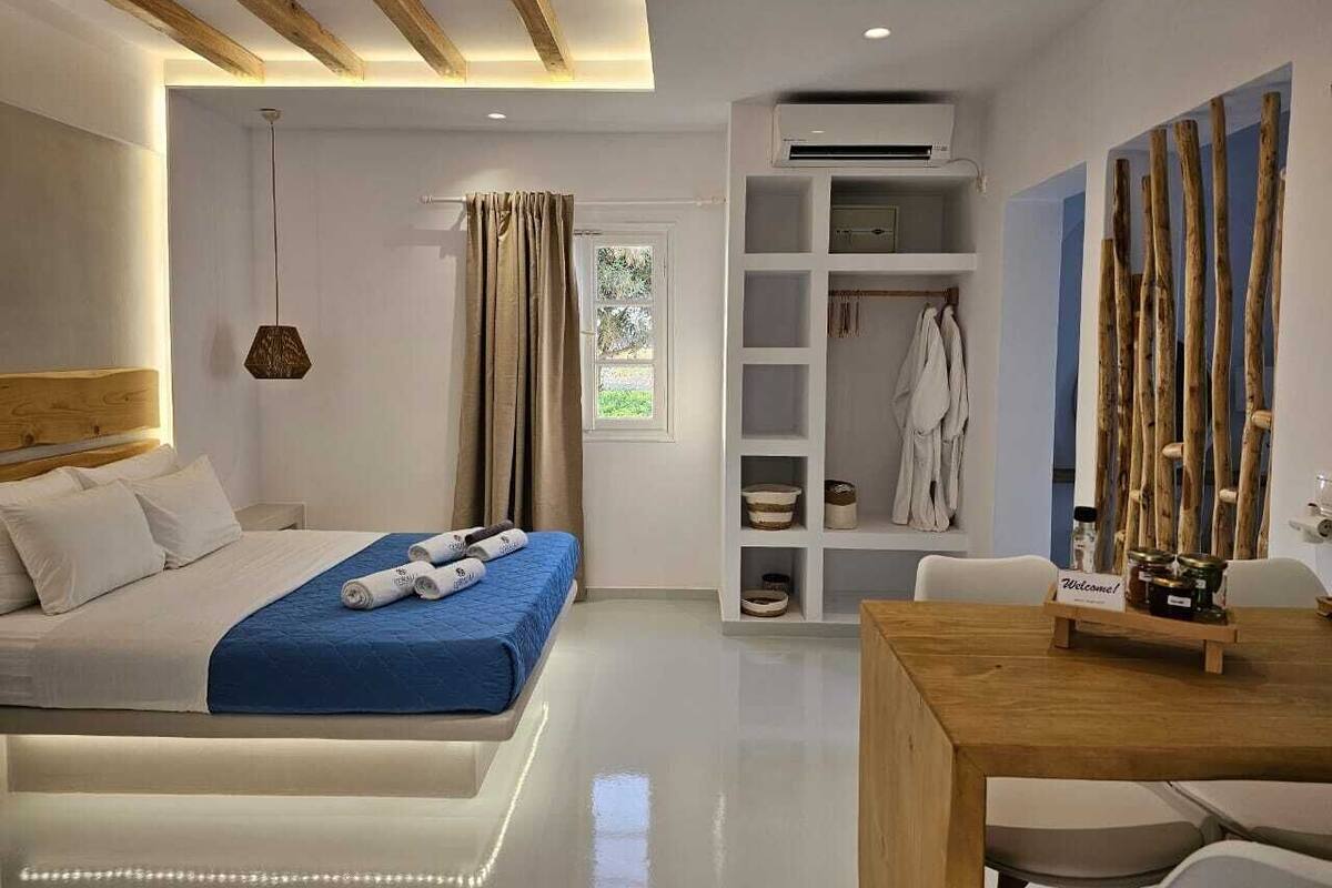A modern hotel room with a bed, a wooden dining table, a built-in wardrobe, and a large window with curtains.