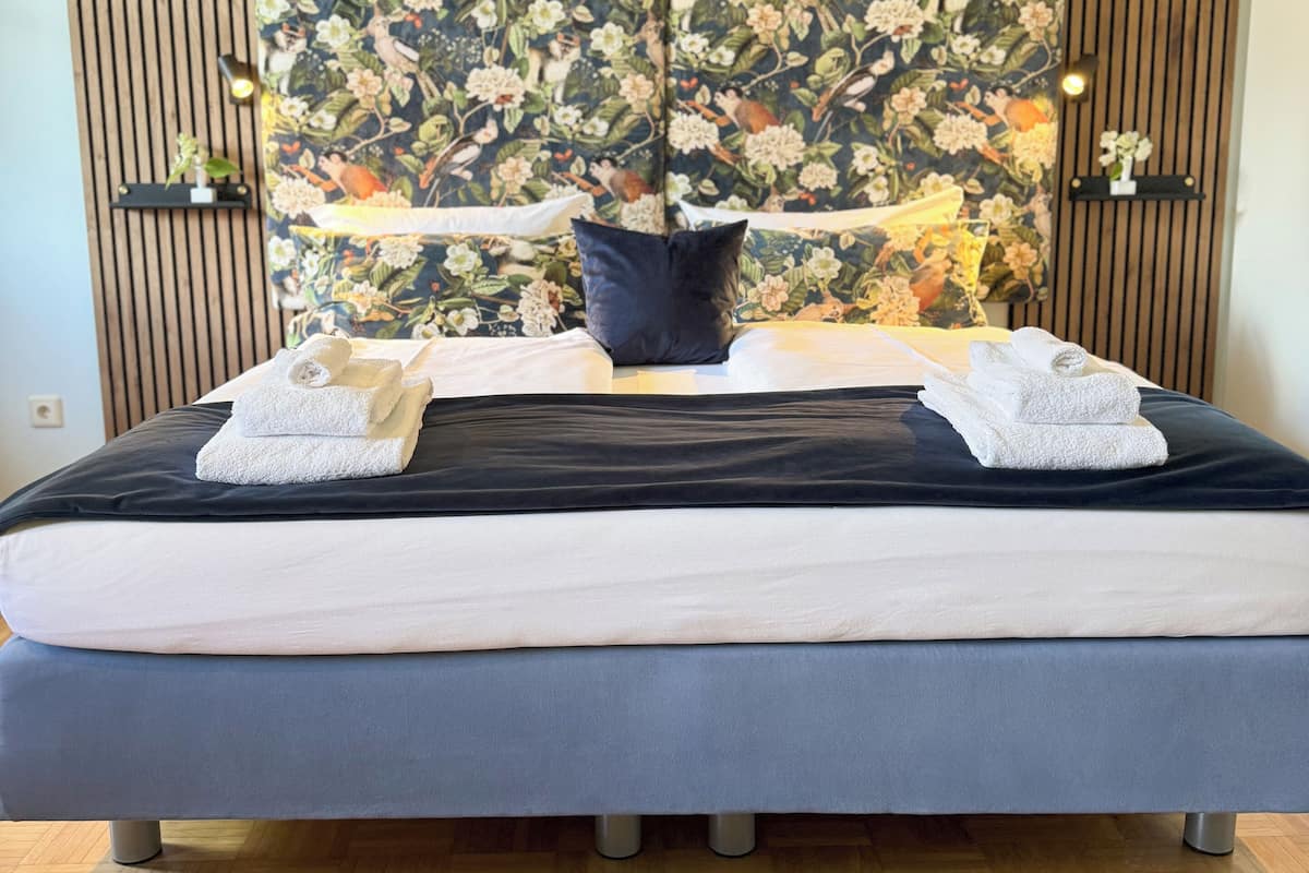A neatly made bed with white and dark blue linens, a dark blue pillow, and two folded white towels. The bed is set against a wooden slat wall with a floral tapestry.