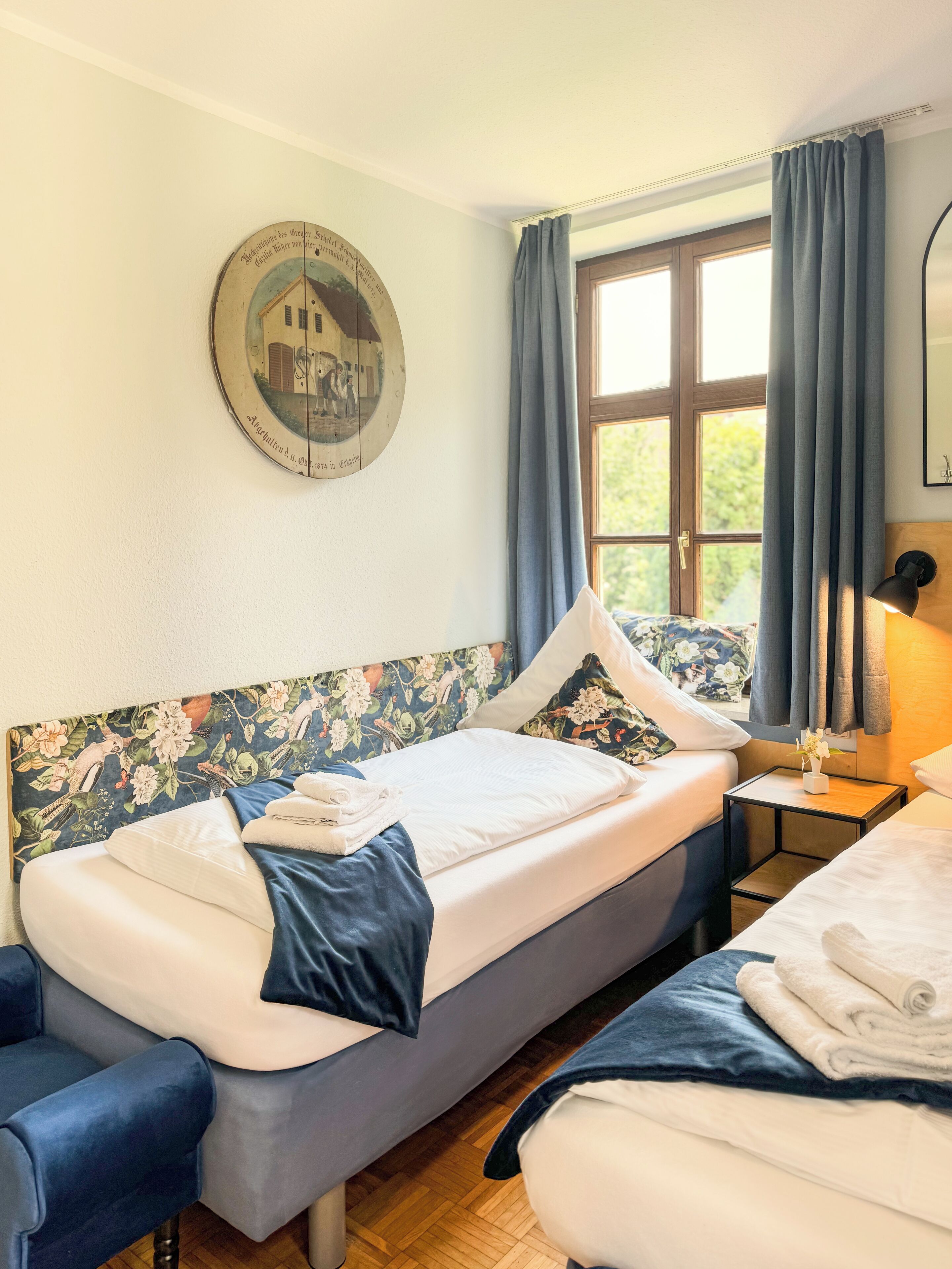 A room with two beds, a wooden headboard with floral patterns, a round wall clock, a window with curtains, and a small table with a lamp.