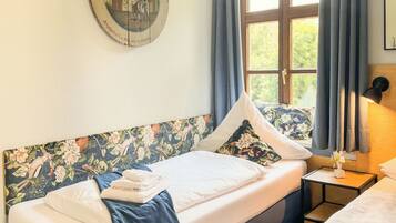 Une chambre avec deux lits, une tête de lit en bois ornée de motifs floraux, une horloge murale ronde, une fenêtre avec des rideaux et une petite table avec une lampe.