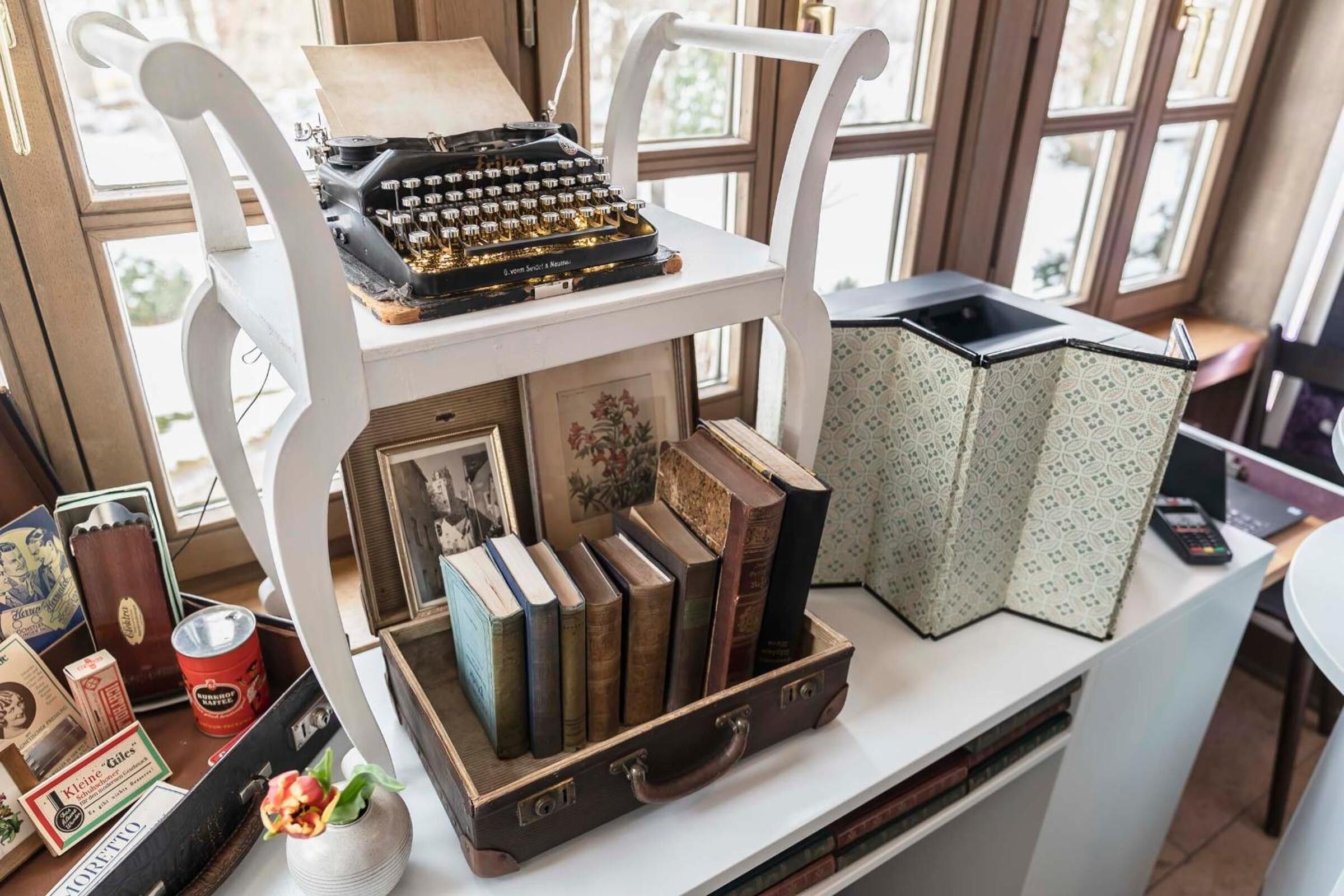 Una máquina de escribir antigua sobre una silla blanca, rodeada de libros y una maleta retro.