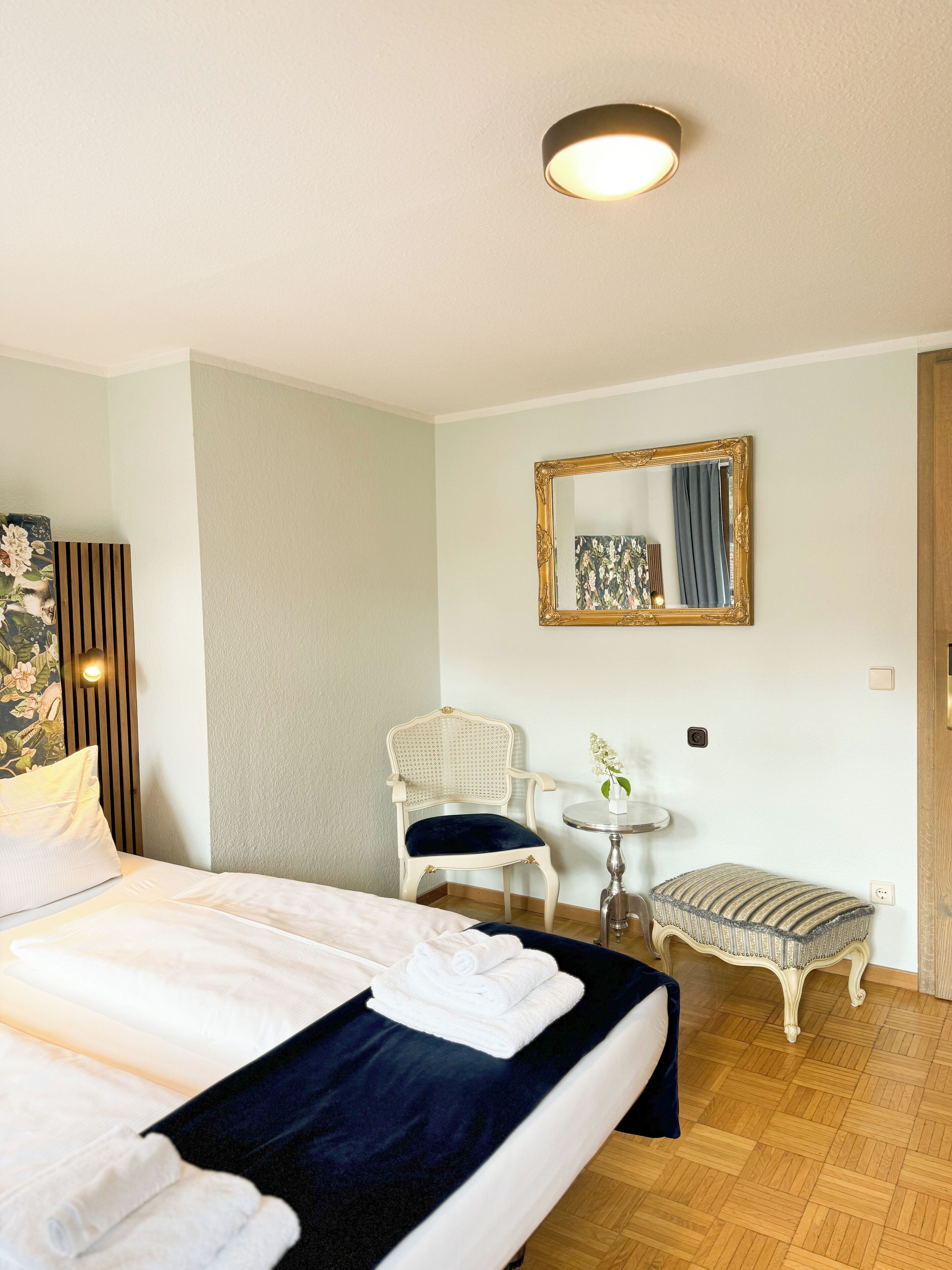 A hotel room with a bed, a chair, a small table with a vase, and a mirror on the wall.