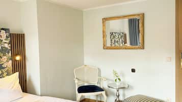 Habitación de hotel con cama, una silla, una mesita con un florero y un espejo en la pared.