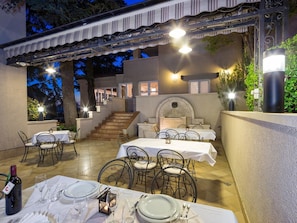 Un espace extérieur aménagé pour les repas, avec des tables pour deux personnes, un escalier menant à un niveau supérieur et une cheminée en pierre.