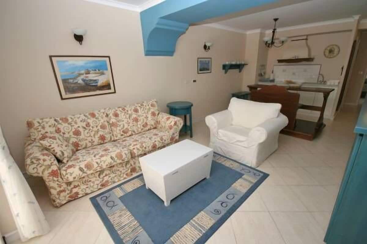 A living room with a floral sofa, a white armchair, and a small white table. There is a painting on the wall and a kitchen area in the background.
