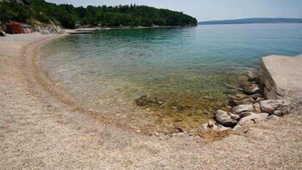 A clear beach with a rocky shoreline, a sandy path leading to the water, and a forested hill in the background.