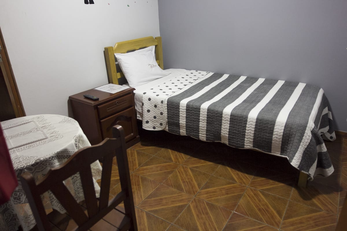 Un dormitorio pequeño y ordenado con una cama individual, una mesita de noche, una silla y una mesa con mantel estampado.