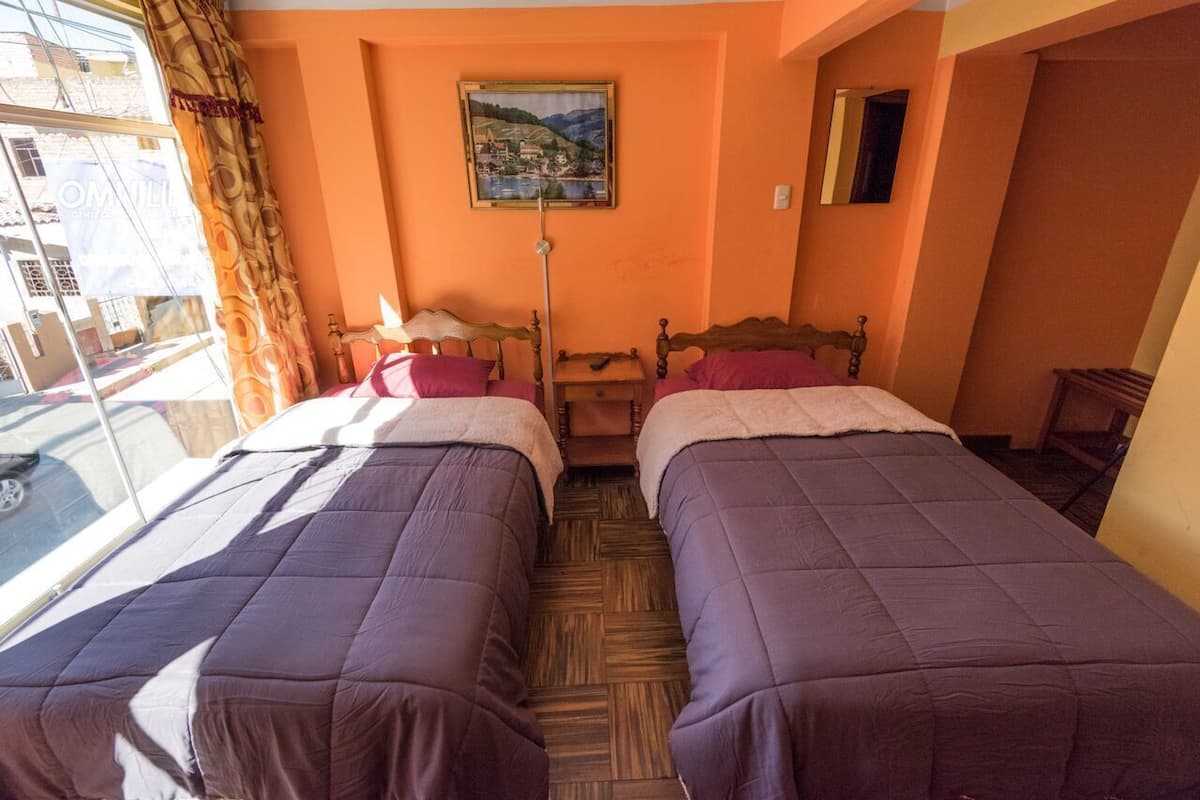 A room with two beds, a window with curtains, a painting on the wall, and a wooden floor.