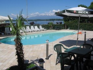 A swimming pool with lounge chairs and a table, surrounded by greenery and a view of a lake. - Hotel Royal (Viverone)
