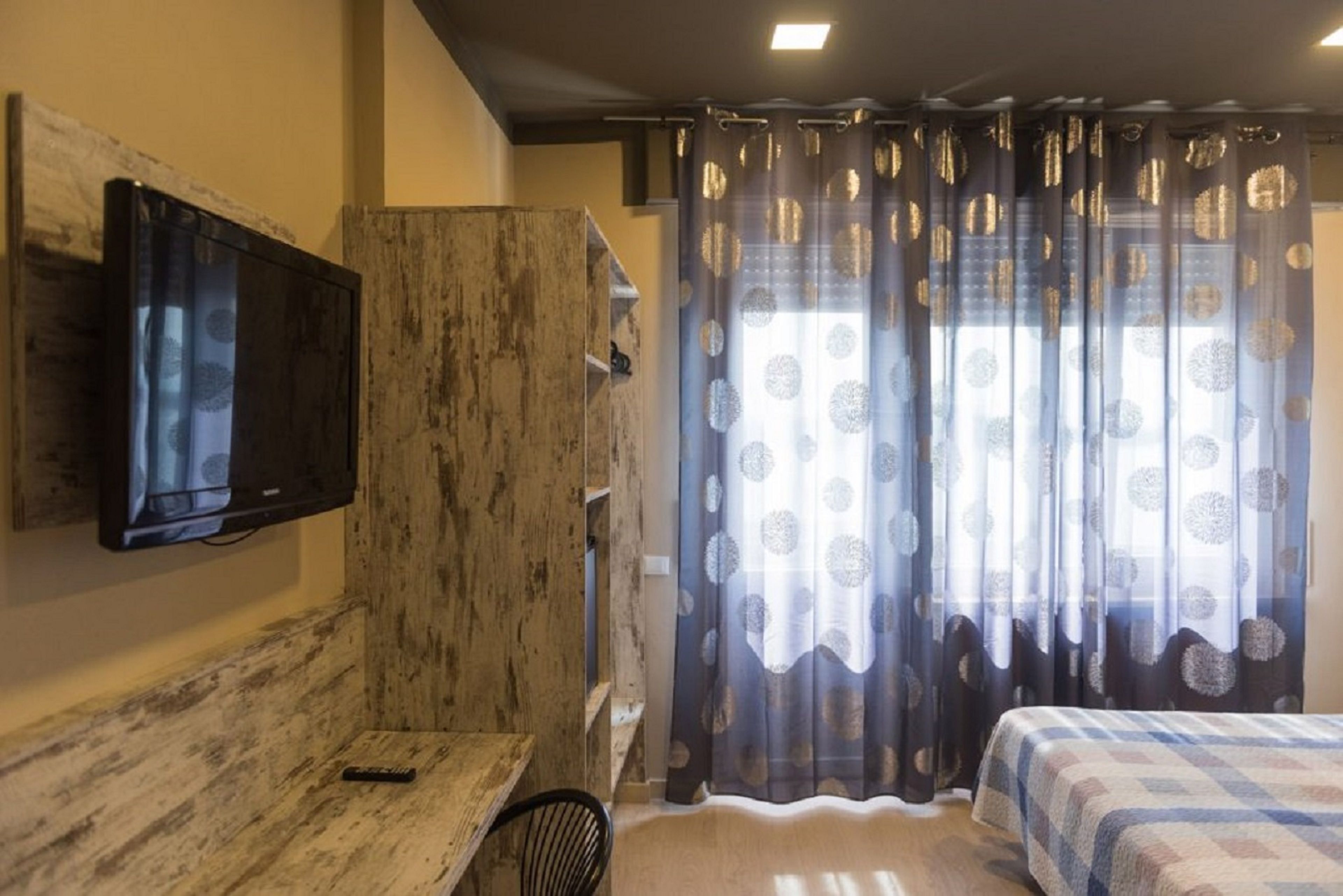 Ein Schlafzimmer mit einem Bett, einem an der Wand befestigten Fernseher, einem Holzschrank und einem Fenster mit gemusterten Vorhängen.