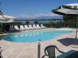 A swimming pool with lounge chairs and an umbrella, surrounded by a tiled deck and a view of a mountain range. - Hotel Royal (Viverone)