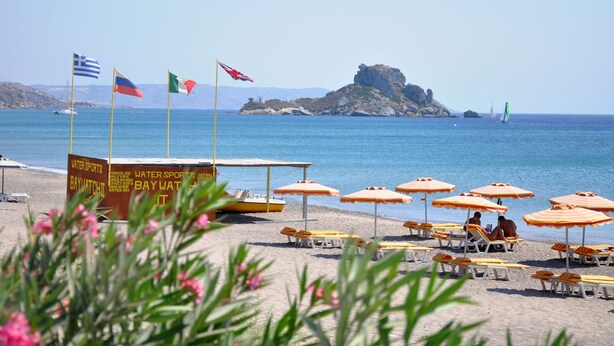 A beach with multiple flags, a water sports sign, and beach umbrellas. - Haralambos Studios (Kos)