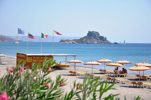 A beach with multiple flags, a water sports sign, and beach umbrellas. - Haralambos Studios (Kos)
