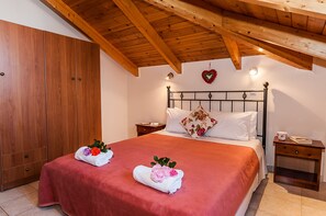 A bedroom with a wooden ceiling, a large wardrobe, a bed with a red bedspread, two towels with flowers, a nightstand, and a heart-shaped decoration on the wall.