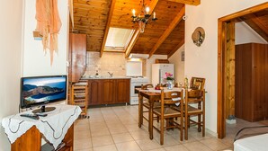 A cozy kitchen with wooden beams, a dining area with a table and chairs, and a TV on a table. - Kavos Psarou Studios and Apartments (Zakynthos)