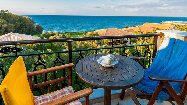 A balcony with a wooden table and chairs, overlooking a coastal view with houses and greenery. - Kavos Psarou Studios and Apartments (Zakynthos)