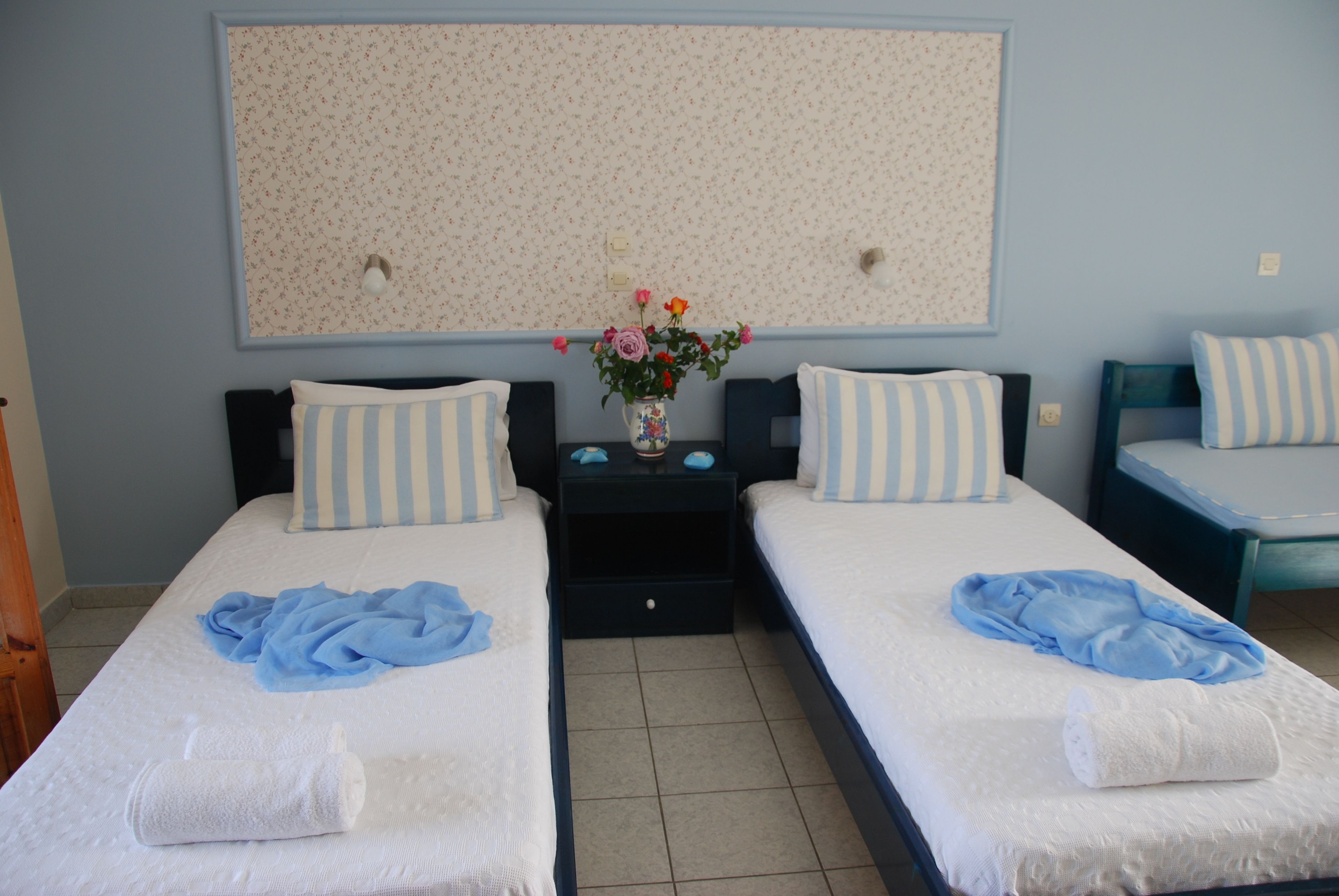 A hotel room with two beds, each with a blue towel and a white towel, a vase with flowers, and a floral-patterned wall hanging.