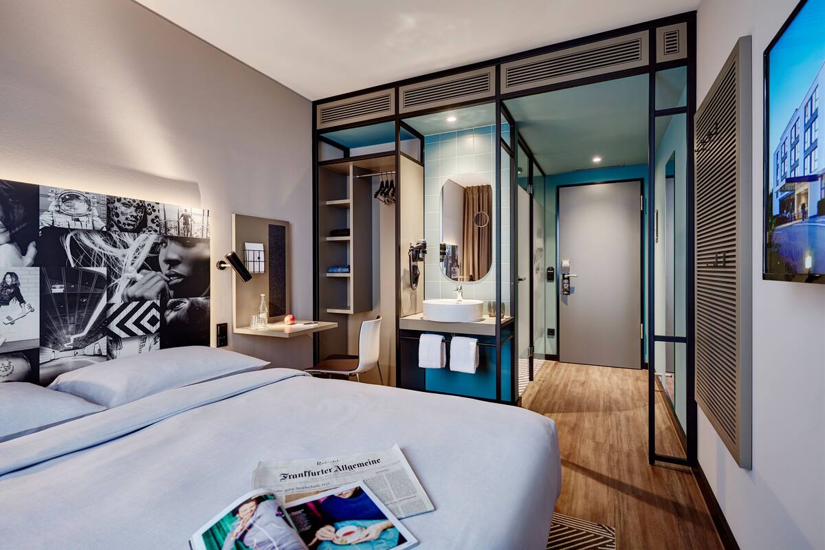 Una habitación de hotel moderna con una cama grande, un baño con espejo y lavamanos, y vistas a la ciudad.