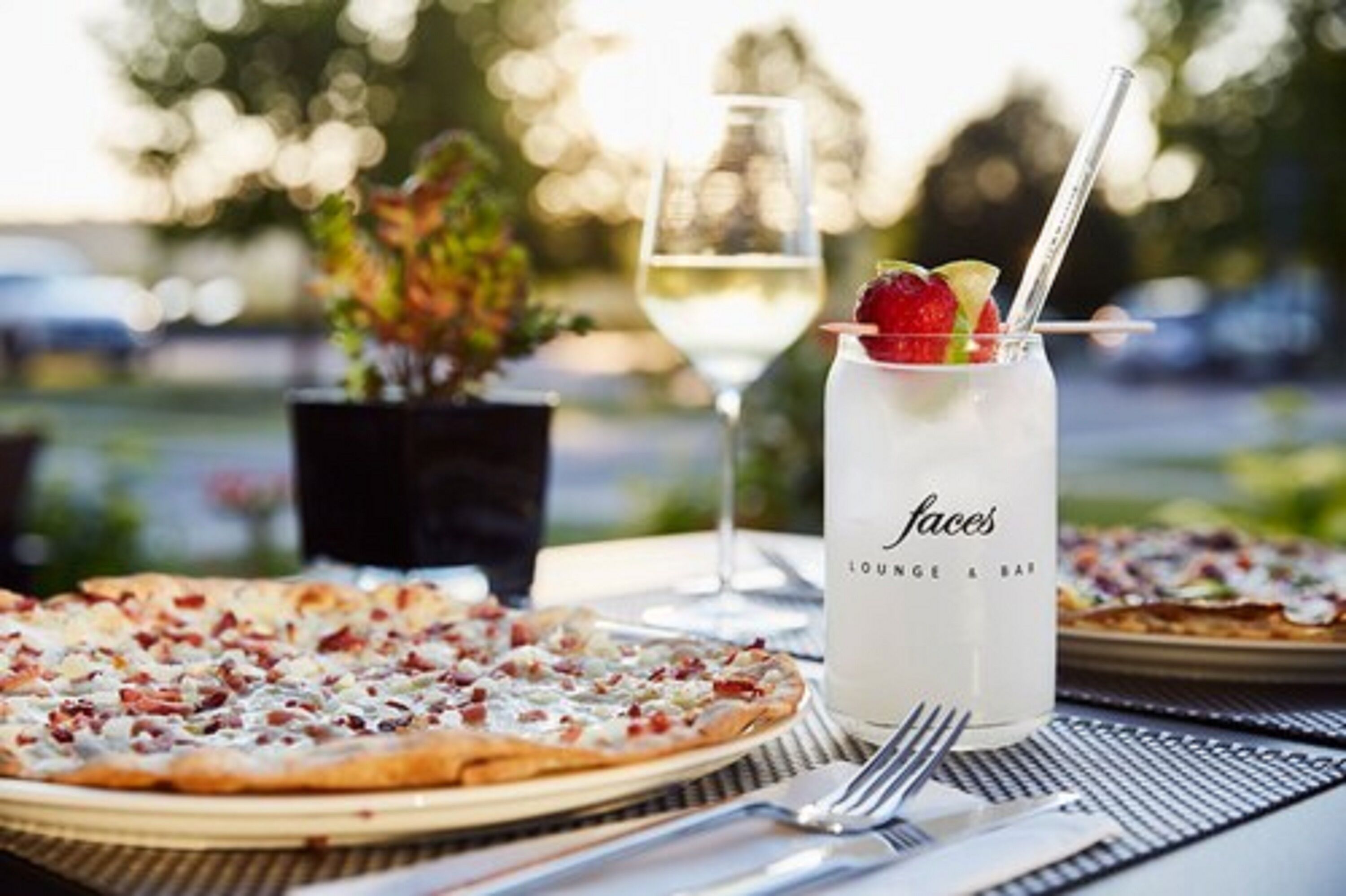 A dining table with a pizza, a glass of white wine, and a cocktail with a straw.