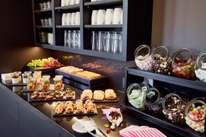 A buffet with various food items and drinks, including pastries, fruits, and jars of candies.