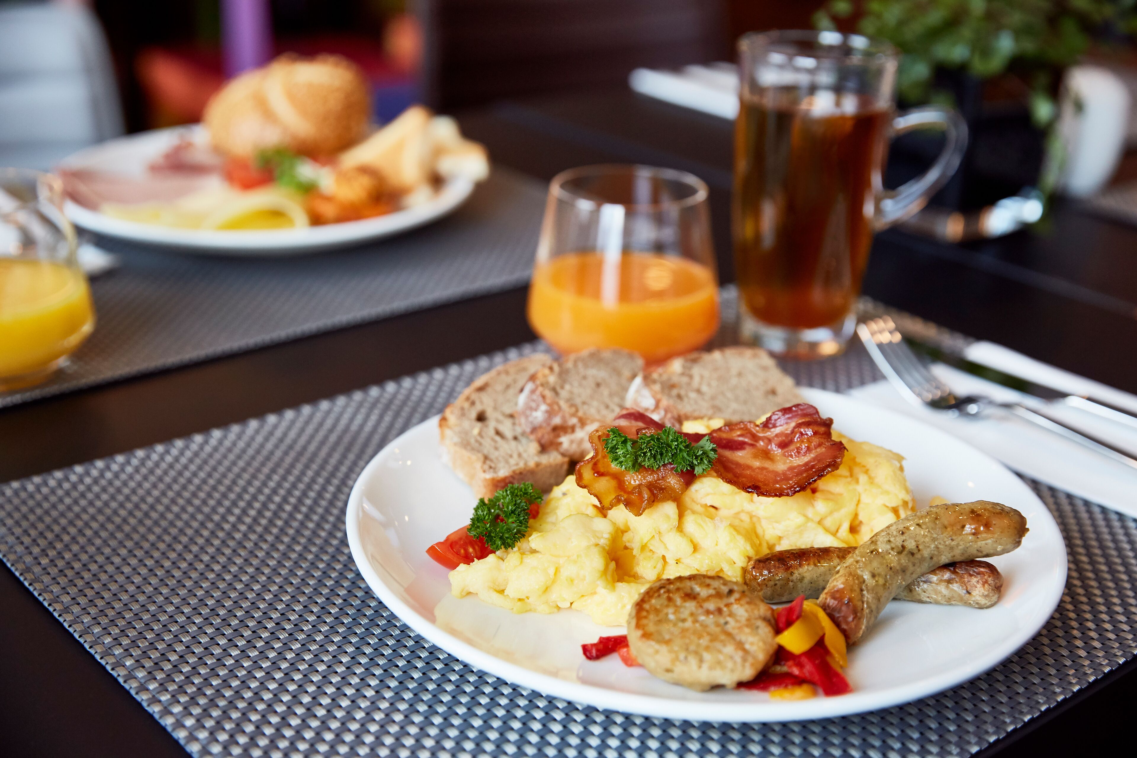 Une assiette de petit-déjeuner comprenant des œufs brouillés, des saucisses et des toasts, accompagnée d’un verre de jus d’orange et d’une tasse de thé.