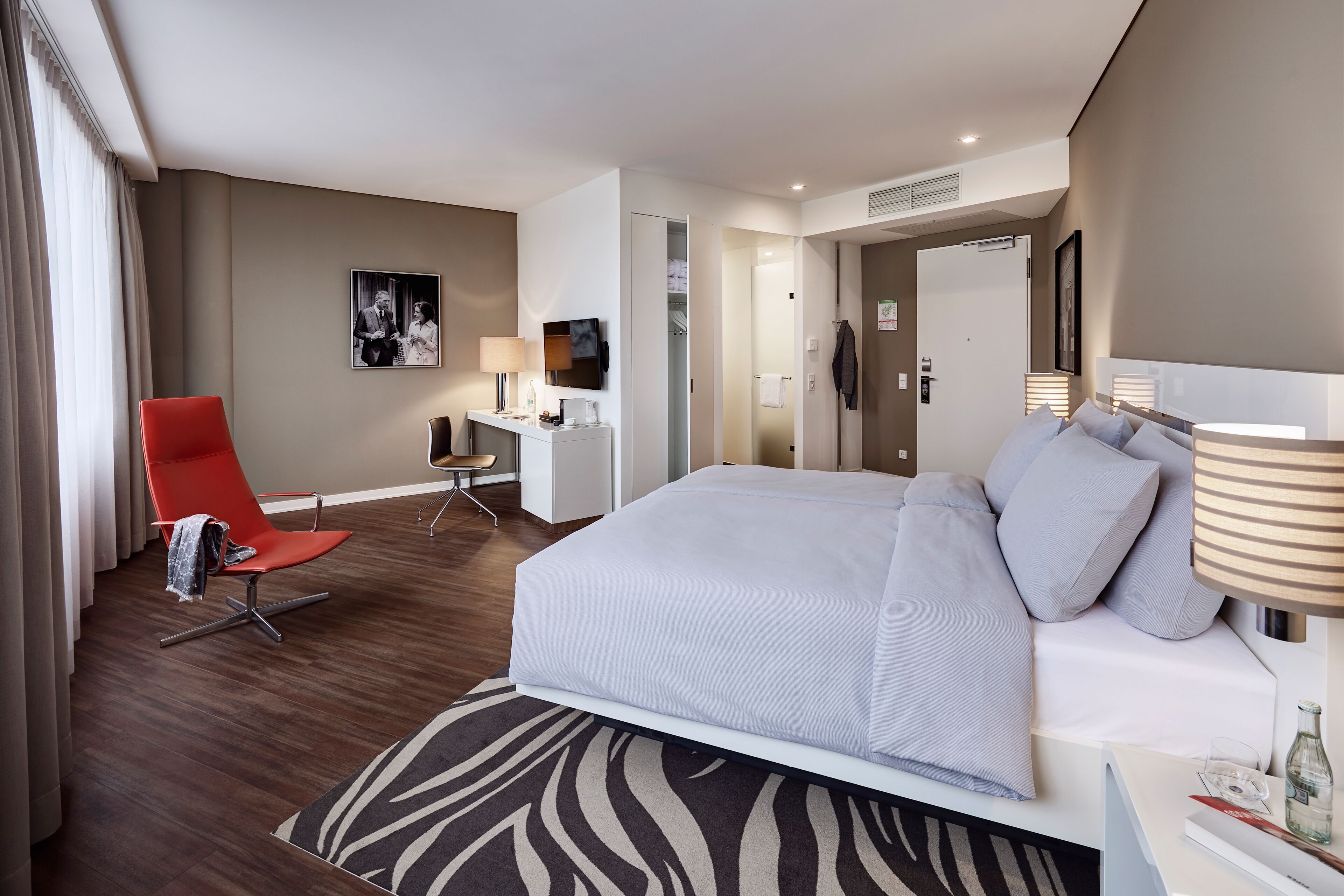 A modern hotel room with a large bed, a desk, a red chair, and a zebra-patterned rug.