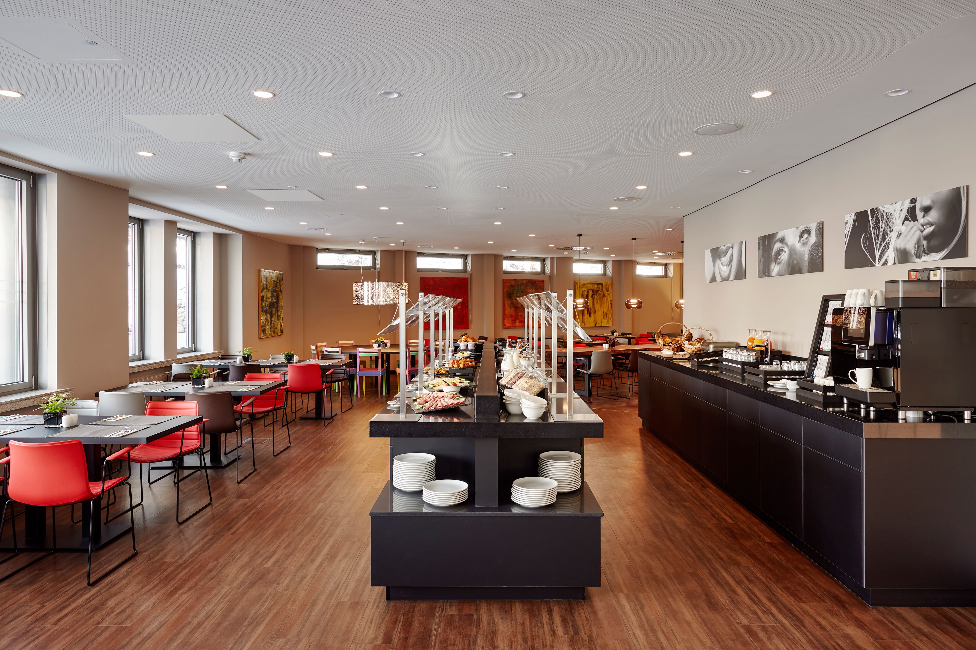 A modern dining area with long tables, red chairs, and a buffet counter with various food items.