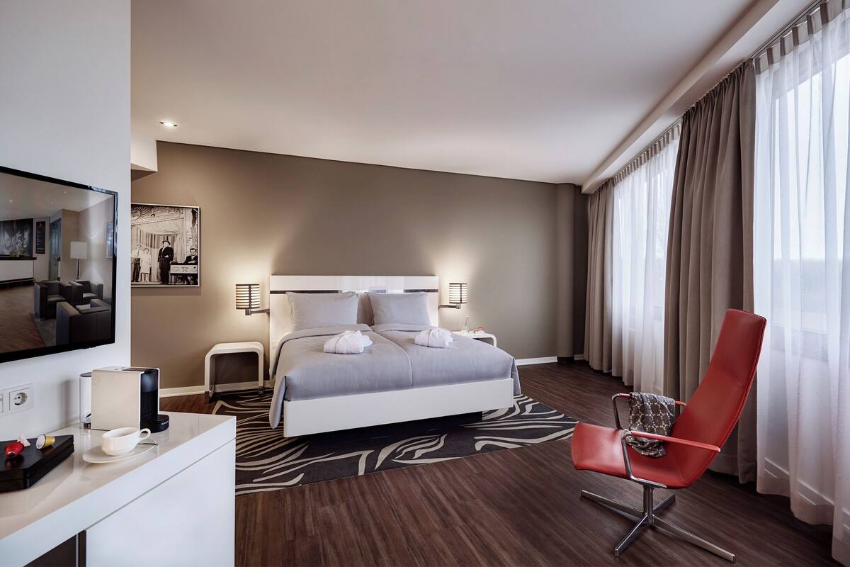 Habitación de hotel moderna con una cama grande, un sillón rojo, un televisor de pantalla plana y un escritorio con cafetera.