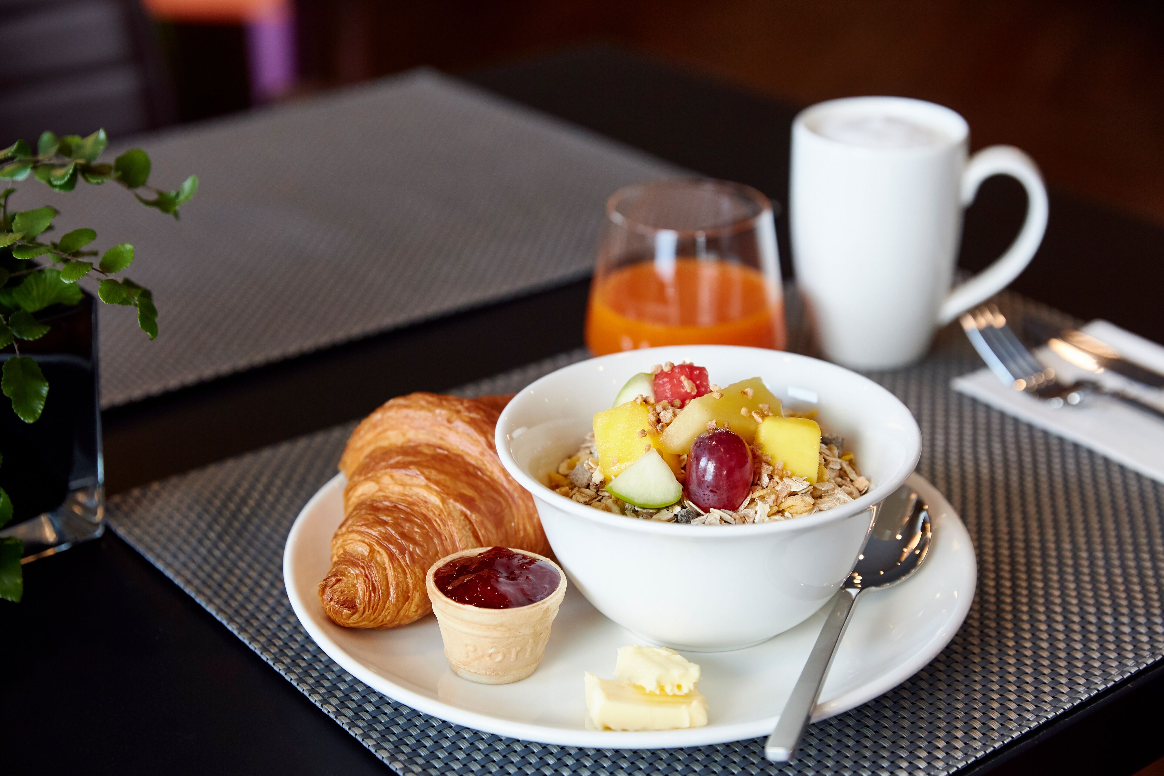 Un desayuno con un croissant, un tazón de cereal con frutas y yogur, un vaso de jugo de naranja y una taza blanca.