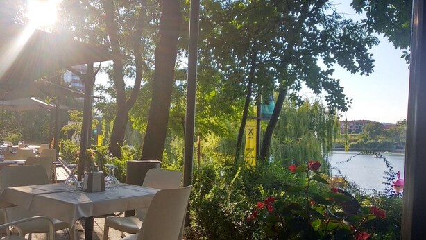An outdoor cafe with tables and chairs, surrounded by greenery and a view of a body of water. -  Univers (T)  (Cluj-Napoca)