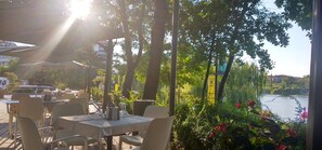 An outdoor cafe with tables and chairs, surrounded by greenery and a view of a body of water.