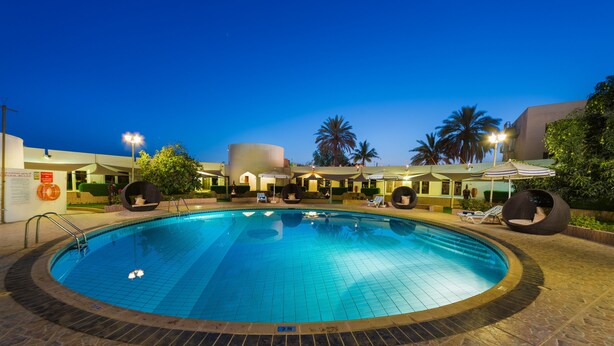 A swimming pool with a tiled deck, surrounded by lounge chairs and umbrellas, with palm trees and a building in the background. - Al Wadi Hotel Sohar (Sohar)