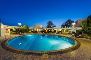 A swimming pool with a tiled deck, surrounded by lounge chairs and umbrellas, with palm trees and a building in the background. - Al Wadi Hotel Sohar (Sohar)