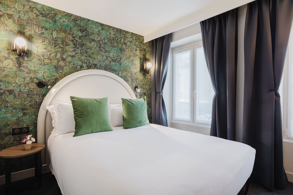 A neatly made bed with white linens and green pillows, a round wooden nightstand with a small vase of flowers, and a wall with a floral pattern.