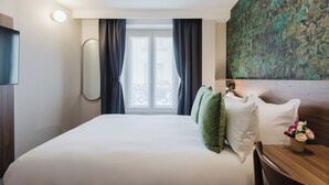 A hotel room with a large bed, a wooden headboard, a small table with a vase of flowers, and a view through a window with curtains. - Prince Albert Opera (Paris)